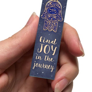 Peut inclure: Un charme de main Hamsa bleu et or avec un &oelig;il bleu et le mot h&eacute;breu pour "protection" au centre. Le charme est sur un fond bleu fonc&eacute; avec des &eacute;toiles blanches et le texte "Trouvez la JOIE dans le voyage".