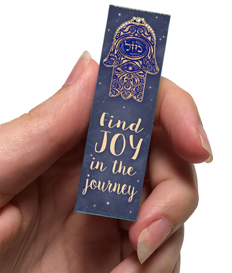 Peut inclure: Un charme de main Hamsa bleu et or avec un &oelig;il bleu et le mot h&eacute;breu pour "protection" au centre. Le charme est sur un fond bleu fonc&eacute; avec des &eacute;toiles blanches et le texte "Trouvez la JOIE dans le voyage".