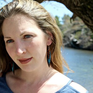 May include: A woman with long blonde hair wearing a blue shirt and turquoise and brown feather earrings.