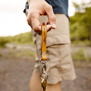 May include: A brown leather keychain with the word "AUSTIN" engraved on it. The keychain has a silver key ring and a silver clip. The keychain is being held by a person's hand.