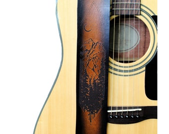 May include: A brown and black leather guitar strap with a mountain and forest design. The strap is attached to a natural wood acoustic guitar. The design includes a triangle, crescent moon, and detailed mountain and tree imagery.