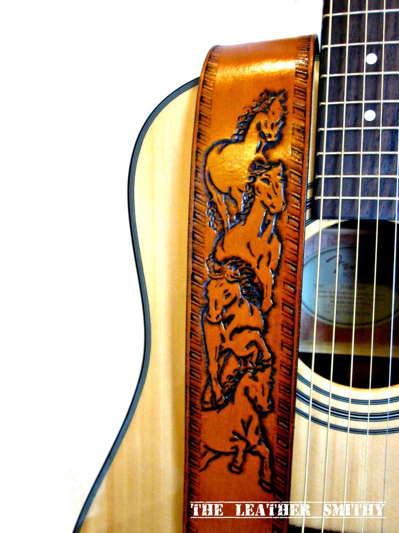 May include: A brown leather guitar strap with a carved design of several horses. The strap is attached to a light-colored acoustic guitar. The text "THE LEATHER SMITHY" is visible at the bottom of the strap.