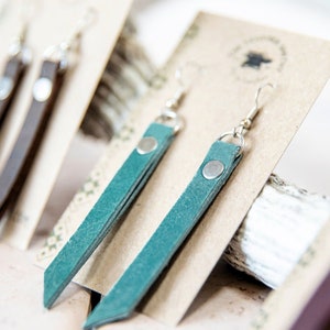 May include: Two pairs of leather earrings, one pair is brown and the other is teal. The earrings are displayed on a card with a small logo.