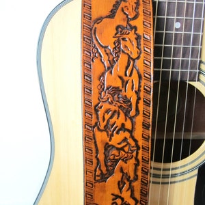 May include: A light brown leather guitar strap with a carved design of horses. The strap is positioned next to a light-colored acoustic guitar. The text "THE LEATHER SMITH" is at the bottom of the strap.