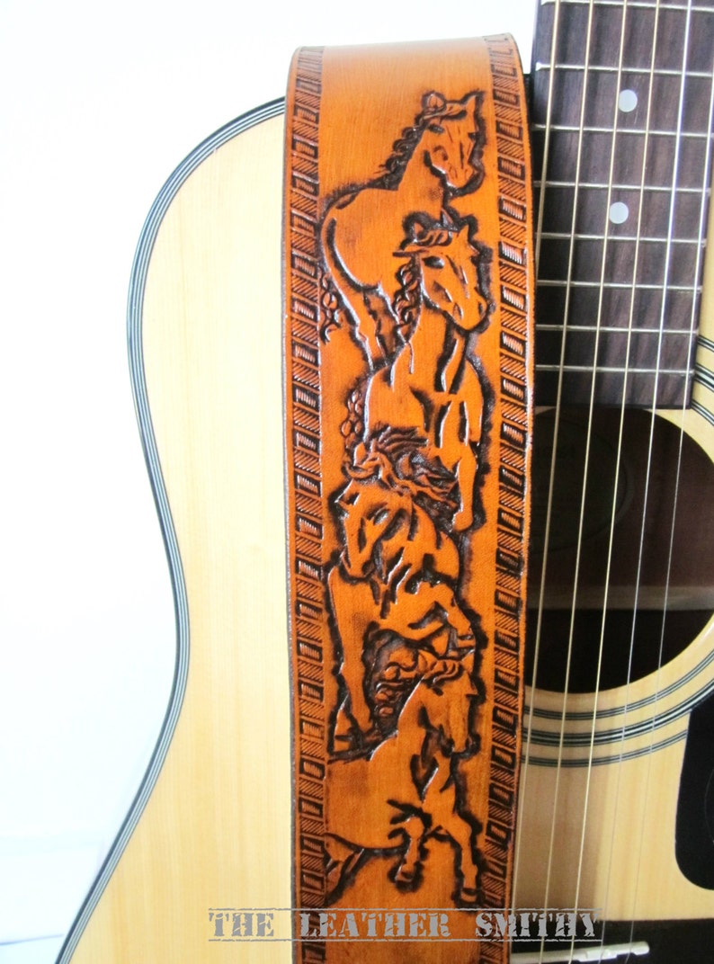 May include: A light brown leather guitar strap with a carved design of horses. The strap is positioned next to a light-colored acoustic guitar. The text "THE LEATHER SMITH" is at the bottom of the strap.