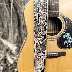 May include: Acoustic guitar with a decorative leather guitar strap. The strap is black with a sunflower design in a lighter color. The guitar is light brown with a dark brown sound hole.