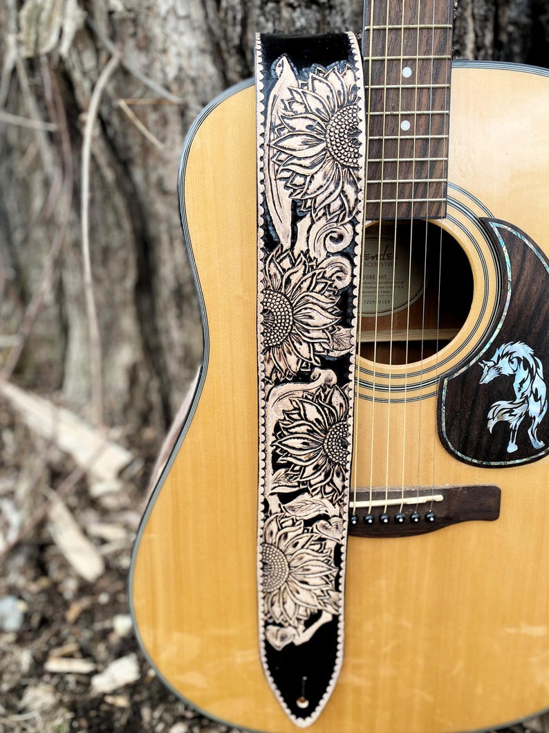 May include: Acoustic guitar with a decorative leather guitar strap. The strap is black with a sunflower design in a lighter color. The guitar is light brown with a dark brown sound hole.