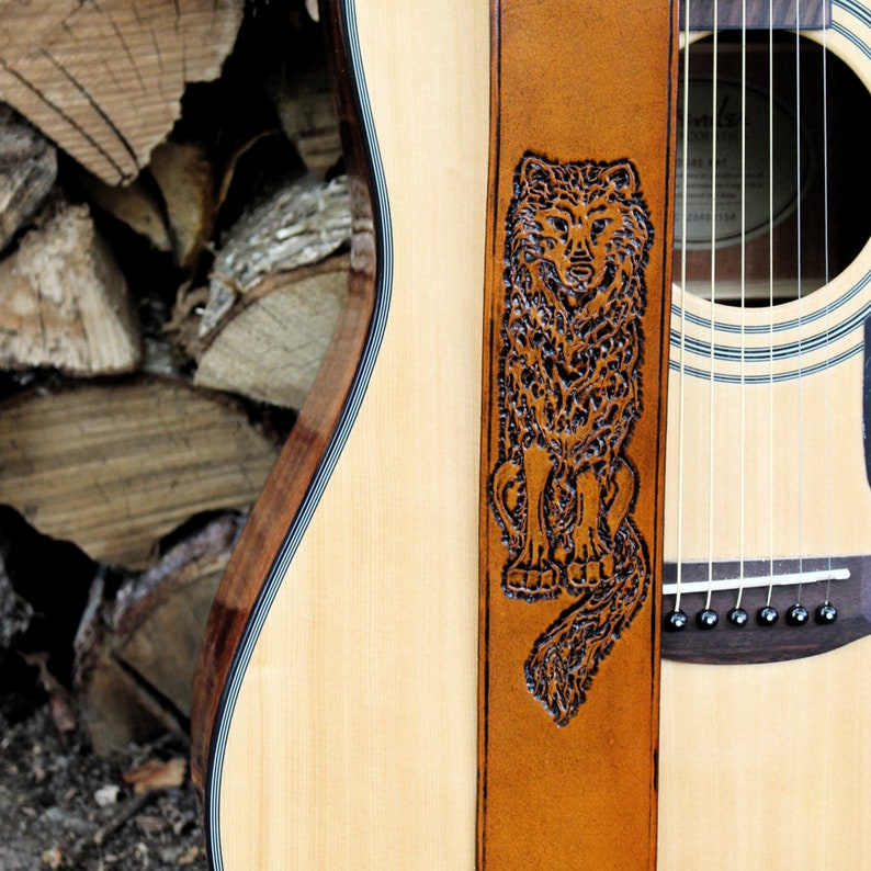 Może przedstawiać: Gitara akustyczna z brązowym sk&oacute;rzanym paskiem z szczeg&oacute;łowym rzeźbionym wzorem wilka. Pasek jest przymocowany do korpusu gitary, prezentując usłojenie drewna i struny instrumentu. Tło obejmuje stos drewna opałowego.