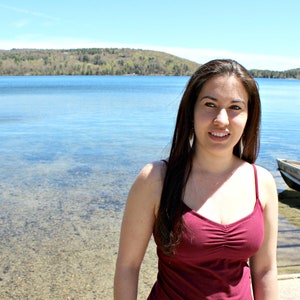 May include: A woman with long brown hair is smiling at the camera. She is wearing a red tank top and is standing in front of a lake.