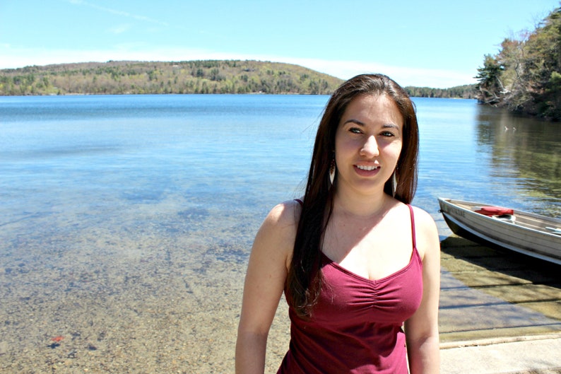 May include: A woman with long brown hair is smiling at the camera. She is wearing a red tank top and is standing in front of a lake.