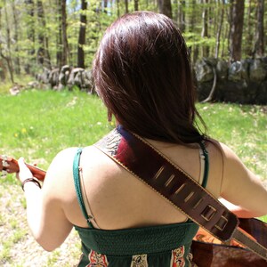 May include: Acoustic guitar with a brown leather strap, worn over the shoulder. The strap has decorative stitching and a patterned design. The person is wearing a green dress. The background is a natural outdoor setting with trees and grass.