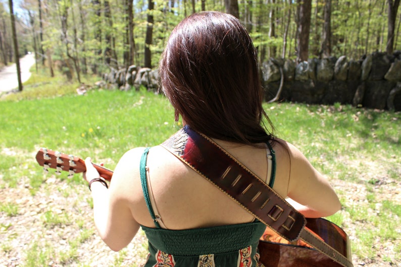 May include: Acoustic guitar with a brown leather strap, worn over the shoulder. The strap has decorative stitching and a patterned design. The person is wearing a green dress. The background is a natural outdoor setting with trees and grass.