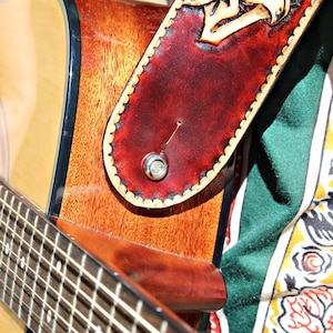 May include: Close-up of an acoustic guitar with a brown wooden body and a fretboard. A burgundy leather guitar strap with a carved sunflower design is attached. The strap has a scalloped edge and a silver button.