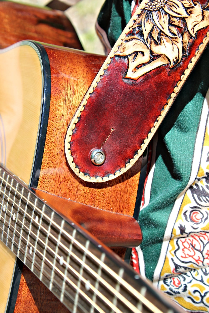 May include: Close-up of an acoustic guitar with a brown wooden body and a fretboard. A burgundy leather guitar strap with a carved sunflower design is attached. The strap has a scalloped edge and a silver button.