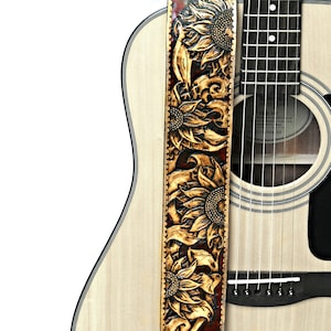 May include: Acoustic guitar with a custom leather guitar strap. The strap is tan with a dark brown border and features a carved sunflower design. The guitar has a natural wood finish.