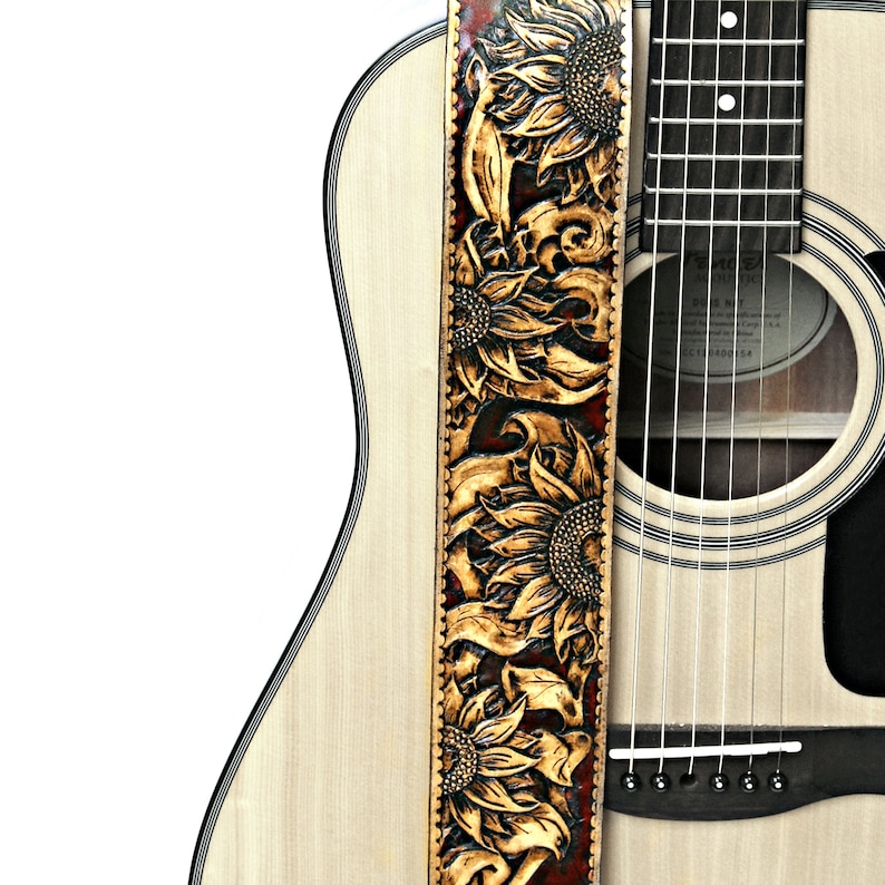 May include: Acoustic guitar with a custom leather guitar strap. The strap is tan with a dark brown border and features a carved sunflower design. The guitar has a natural wood finish.