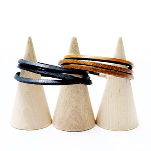 Puede incluir: Dos pulseras de cuero, una negra y otra marrón, expuestas en soportes de madera en forma de cono.
