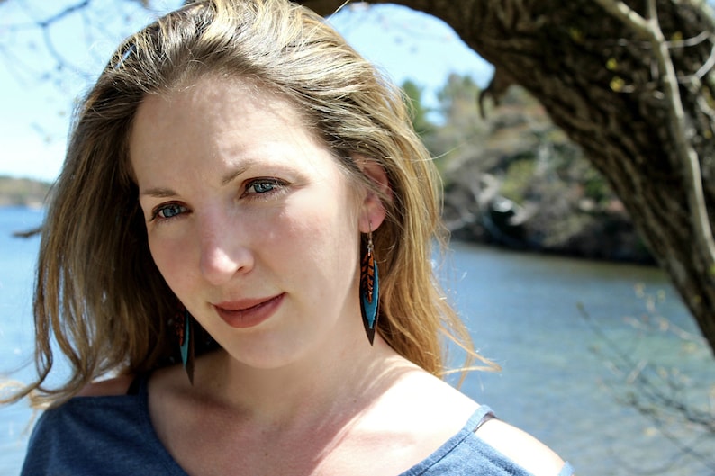 May include: A woman with long blonde hair wearing a blue shirt and turquoise and brown feather earrings.