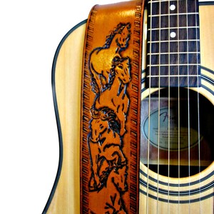 May include: A brown leather guitar strap with a carved design of several horses. The strap is attached to a natural wood acoustic guitar. The text "THE LEATHER SMITHY" is at the bottom of the strap.