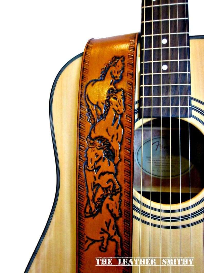 May include: A brown leather guitar strap with a carved design of several horses. The strap is attached to a natural wood acoustic guitar. The text "THE LEATHER SMITHY" is at the bottom of the strap.