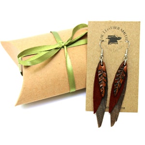 May include: A pair of leather earrings with a leaf design. The earrings are brown and have a silver hook. The earrings are packaged in a brown box with a green ribbon. The box has a label that says "The Leather Smith, Art in Leather".