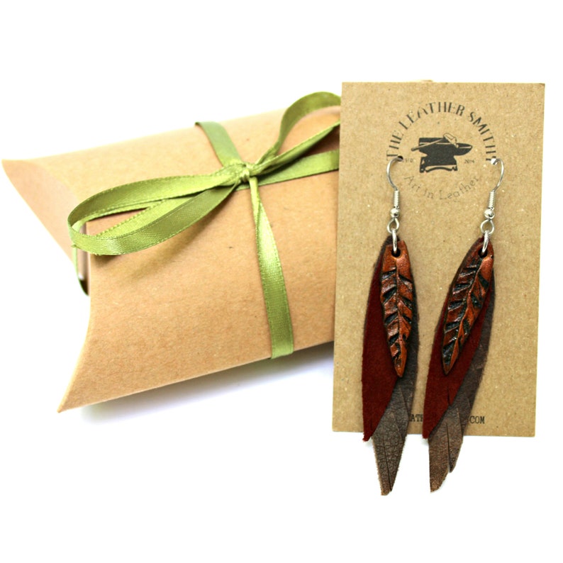 May include: A pair of leather earrings with a leaf design. The earrings are brown and have a silver hook. The earrings are packaged in a brown box with a green ribbon. The box has a label that says "The Leather Smith, Art in Leather".