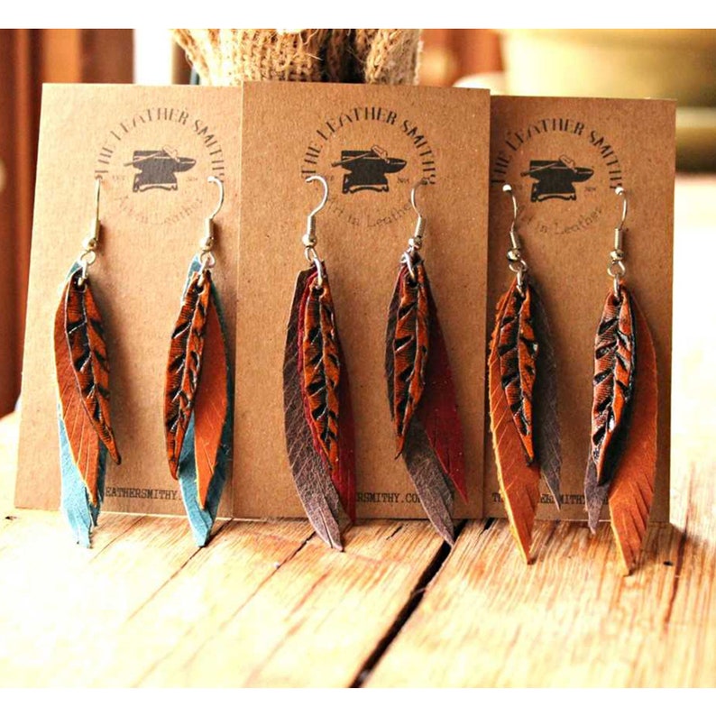 May include: Three pairs of leather feather earrings in brown, teal, and burgundy. The earrings are handmade and feature a layered design. The earrings are displayed on brown cards with the text "The Leather Smith" printed on them.