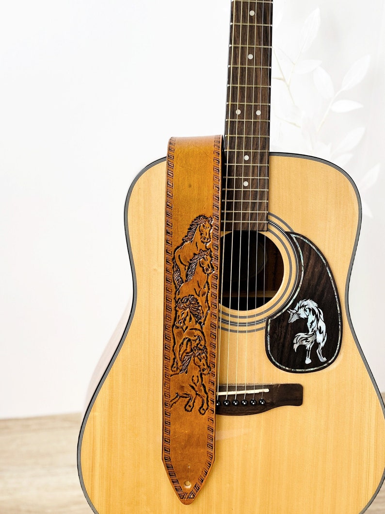 May include: Acoustic guitar with a brown leather strap featuring a carved horse design. The guitar has a natural wood finish and a decorative pickguard with a wolf design.