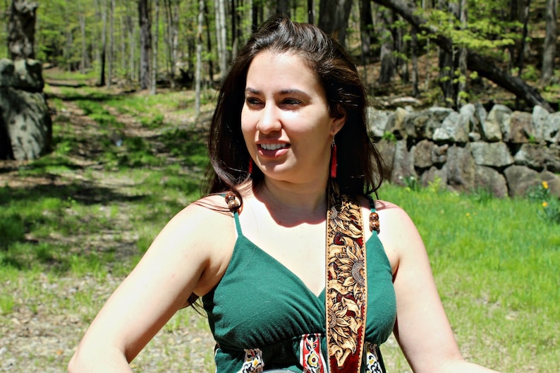May include: A woman wearing a green spaghetti strap dress with a decorative strap featuring a sunflower design. She is also wearing red earrings. The background includes a stone wall, trees, and green grass.