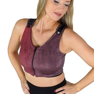 Puede incluir: Primer plano de una mujer con un sujetador deportivo morado y rojo con una cremallera en la parte delantera. El sujetador tiene una banda negra en la parte inferior y tirantes negros. La mujer lleva pantalones negros.