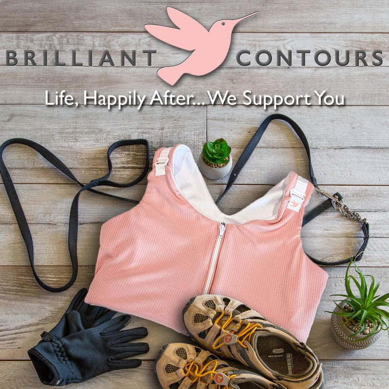 May include: A pink sports bra with a zipper closure and adjustable straps. The bra is on a wooden surface with a pair of hiking boots, gloves, and a leash.