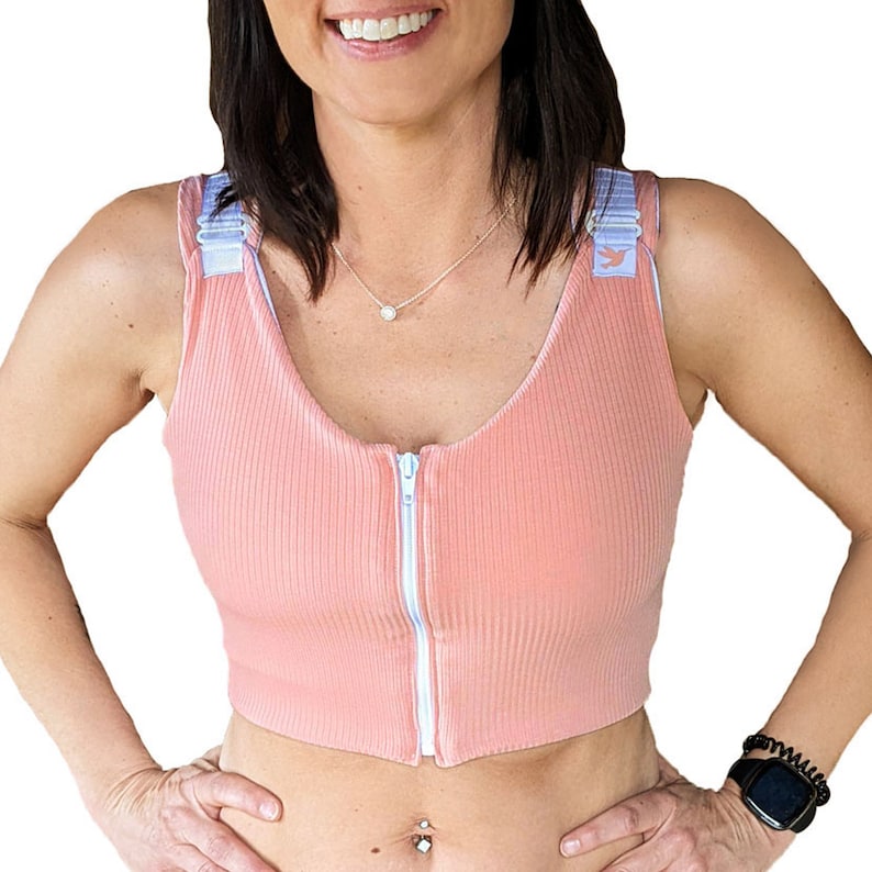 May include: A pink, ribbed, front-zip sports bra with adjustable straps and a white zipper. The straps have a light blue trim and a small bird logo.