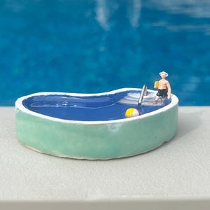 Könnte beinhalten: Miniatur-Keramikbecken mit hellgrüner Außenseite und blauem Innenraum. Eine kleine Figur mit Hut und Shorts steht im Becken und hält ein Getränk. Ein gelb-weißer Strandball schwimmt in der Nähe. Der Hintergrund ist ein verschwommenes Blau.