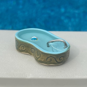 May include: A miniature ceramic bathtub with a light blue interior and a silver-colored handle. The exterior features a green and brown swirl design. A small, blue bubble floats in the water.