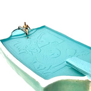 May include: A miniature turquoise swimming pool with a diving board and a person sitting on the edge. The pool has a white border and a fish design on the bottom. The person is wearing black swim trunks.