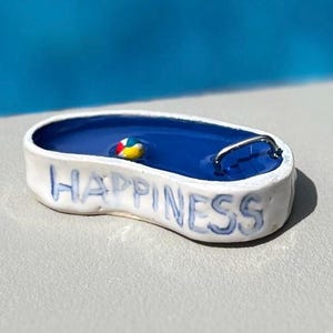 May include: A miniature ceramic pool with blue water and a small, colorful beach ball. The word "HAPPINESS" is written in blue on the white exterior. A silver metal handle is attached to the side of the pool.