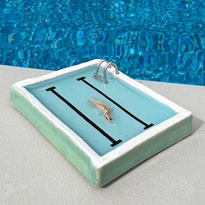 Puede incluir: Una piscina en miniatura con un nadador. La piscina tiene un interior azul claro, líneas negras y una escalera plateada. El borde de la piscina es verde claro con un borde blanco. El fondo es una piscina más grande.