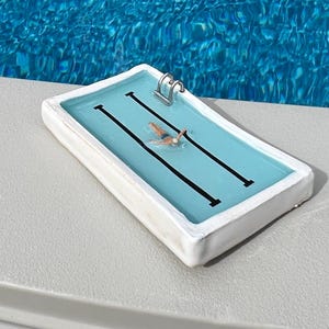 Puede incluir: Una piscina rectangular en miniatura con un nadador. La piscina tiene un interior azul claro con líneas de carrera negras y una escalera plateada. La piscina está sobre una superficie gris, con una piscina más grande en el fondo.
