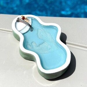 May include: A miniature swimming pool charm with a light blue interior, white trim, and a green exterior. A tiny figure is sunbathing near the edge. The charm has a silver-coloured loop for attaching to a keychain or necklace.