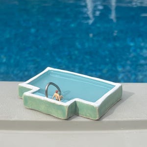 Könnte beinhalten: Ein Miniatur-Keramikbecken mit hellgrüner Außenseite und hellblauem Innenraum. Eine winzige Figur befindet sich im Becken, mit einer silbernen Metallstange. Das Becken befindet sich auf einer grauen Oberfläche, mit einem blauen Becken im Hintergrund.