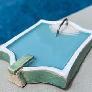 Può includere: Statuetta di piscina in miniatura con interno azzurro, bordi bianchi e trampolino verde. Una maniglia in metallo argentato è attaccata alla sezione bianca. La statuetta è ambientata su uno sfondo blu sfocato.