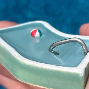 Könnte beinhalten: Ein Miniatur-Keramikbecken mit hellblauem Innenraum und einem kleinen, bunten Strandball, der im Wasser schwimmt. Eine silberfarbene Metallleiter ist an der Seite des Beckens befestigt. Das Becken hat einen hellgrünen Rand.