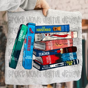 May include: A gray sweatshirt featuring a stack of books with titles like "The Housemaid" and "The Coworker." The books have colorful covers and the author's name, Freida McFadden, is visible. The words "Broke Moms digital designs" are printed on the sweatshirt.