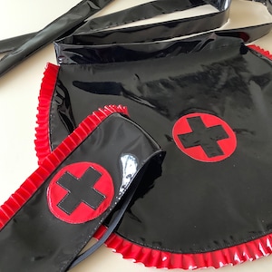 Op de afbeelding: Zwart vinyl verpleegster outfit met rode kruis emblemen. De outfit bevat een schort met een gerimpelde rand, een hoofdband en strikken. De rode kruizen staan op een rode cirkel. Glanzende afwerking.