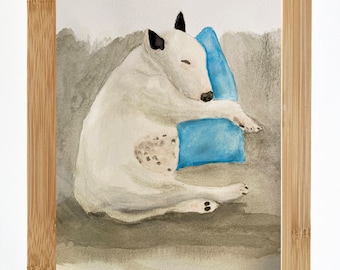 May include: Watercolor painting of a white dog with black ears and nose sleeping on a blue pillow. The dog is curled up on the pillow and has its eyes closed. The painting is framed in a light wood frame. The artist's signature, "T.N.", is in the bottom left corner.