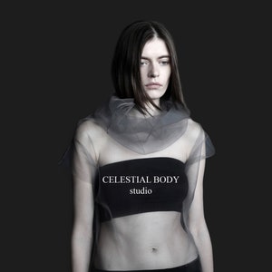 May include: A woman wearing a sheer gray top with a black bandeau underneath. The text "CELESTIAL BODY studio" is printed on the bandeau.