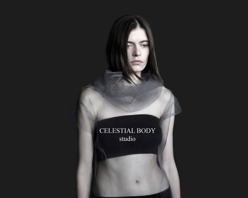 May include: A woman wearing a sheer gray top with a black bandeau underneath. The text "CELESTIAL BODY studio" is printed on the bandeau.