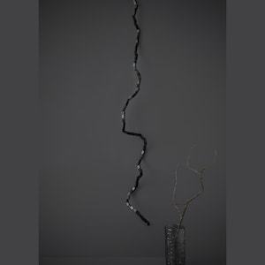 May include: A dark, minimalist composition featuring a long, black, textured decorative element with small, light-colored accents, suspended against a gray backdrop. A vase with branches is at the bottom.