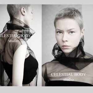 May include: A woman with short, silver hair wears a black sheer top with the text "CELESTIAL BODY studio" on it. The background is a gray wall.