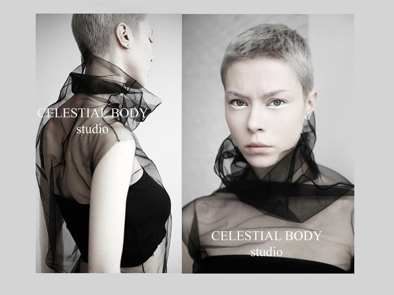 May include: A woman with short, silver hair wears a black sheer top with the text "CELESTIAL BODY studio" on it. The background is a gray wall.